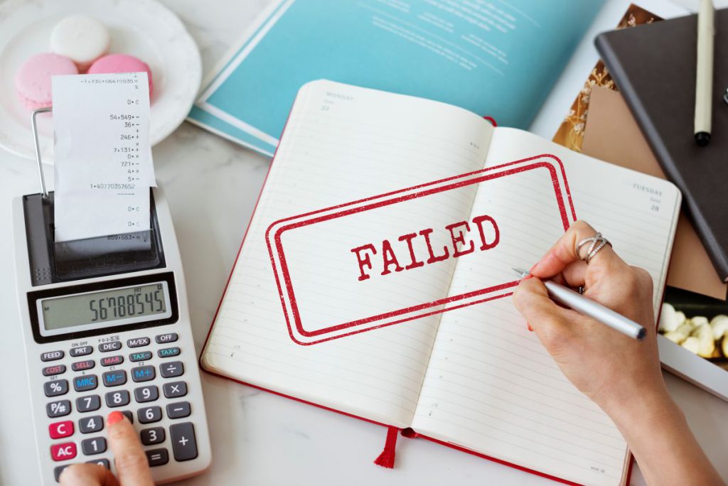 Widok z góry na biurko z kalkulatorem, wydrukiem z bankomatu i otwartym planerem. Na stronach notesu widnieje duża, czerwona pieczątka z napisem „FAILED”, symbolizująca negatywną decyzję kredytową.