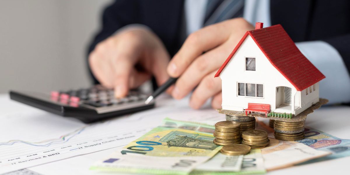 Dłonie osoby w garniturze przy kalkulatorze i wykresach, obok miniaturowego domu stojącego na stosach monet i banknotach euro. Obraz symbolizuje obliczanie rat i planowanie dużego kredytu.