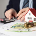 Dłonie osoby w garniturze przy kalkulatorze i wykresach, obok miniaturowego domu stojącego na stosach monet i banknotach euro. Obraz symbolizuje obliczanie rat i planowanie dużego kredytu.