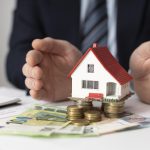 Mężczyzna w garniturze osłania dłońmi miniaturowy model domu z czerwonym dachem, który stoi na stosach monet i banknotach euro.