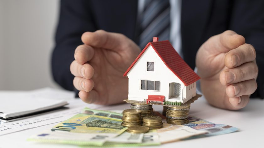 Mężczyzna w garniturze osłania dłońmi miniaturowy model domu z czerwonym dachem, który stoi na stosach monet i banknotach euro.