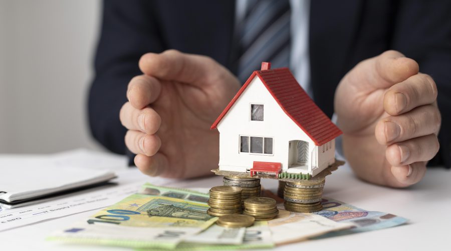 Mężczyzna w garniturze osłania dłońmi miniaturowy model domu z czerwonym dachem, który stoi na stosach monet i banknotach euro.