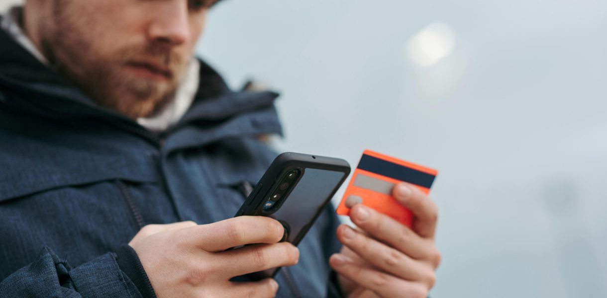 Mężczyzna w niebieskiej kurtce trzymający smartfon i kartę płatniczą, weryfikujący transakcję zbliżeniową.