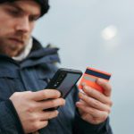 Mężczyzna w niebieskiej kurtce trzymający smartfon i kartę płatniczą, weryfikujący transakcję zbliżeniową.