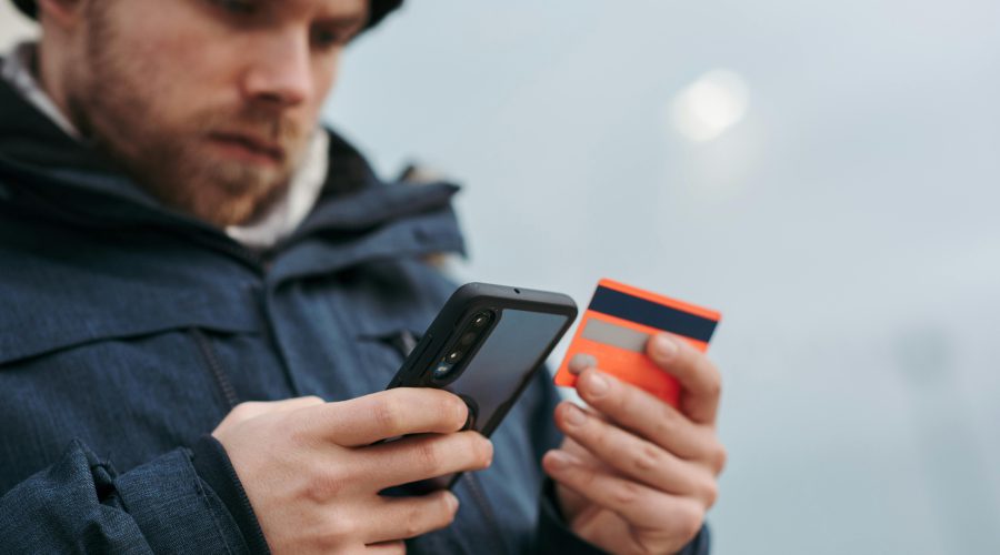 Mężczyzna w niebieskiej kurtce trzymający smartfon i kartę płatniczą, weryfikujący transakcję zbliżeniową.