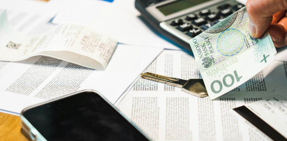 Dłoń trzymająca banknot 100 złotych nad biurkiem, na którym leży czarny kalkulator, klucz, smartfon, długi paragon i dokumenty finansowe.