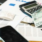 Dłoń trzymająca banknot 100 złotych nad biurkiem, na którym leży czarny kalkulator, klucz, smartfon, długi paragon i dokumenty finansowe.