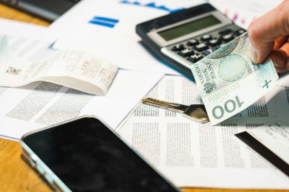 Dłoń trzymająca banknot 100 złotych nad biurkiem, na którym leży czarny kalkulator, klucz, smartfon, długi paragon i dokumenty finansowe.