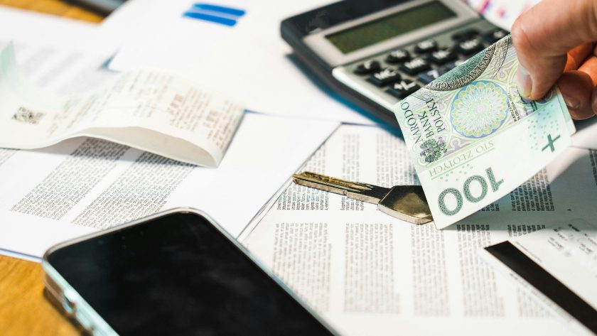 Dłoń trzymająca banknot 100 złotych nad biurkiem, na którym leży czarny kalkulator, klucz, smartfon, długi paragon i dokumenty finansowe.