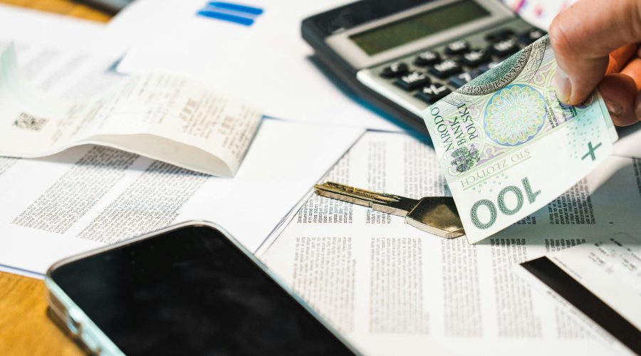 Dłoń trzymająca banknot 100 złotych nad biurkiem, na którym leży czarny kalkulator, klucz, smartfon, długi paragon i dokumenty finansowe.