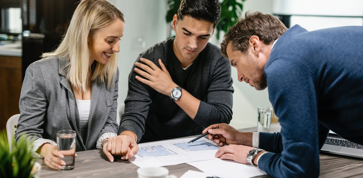 Doradca finansowy wskazuje na dokumenty i omawia plan inwestycyjny z dwojgiem młodych przedsiębiorców przy drewnianym stole.