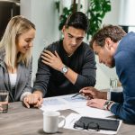 Doradca finansowy wskazuje na dokumenty i omawia plan inwestycyjny z dwojgiem młodych przedsiębiorców przy drewnianym stole.