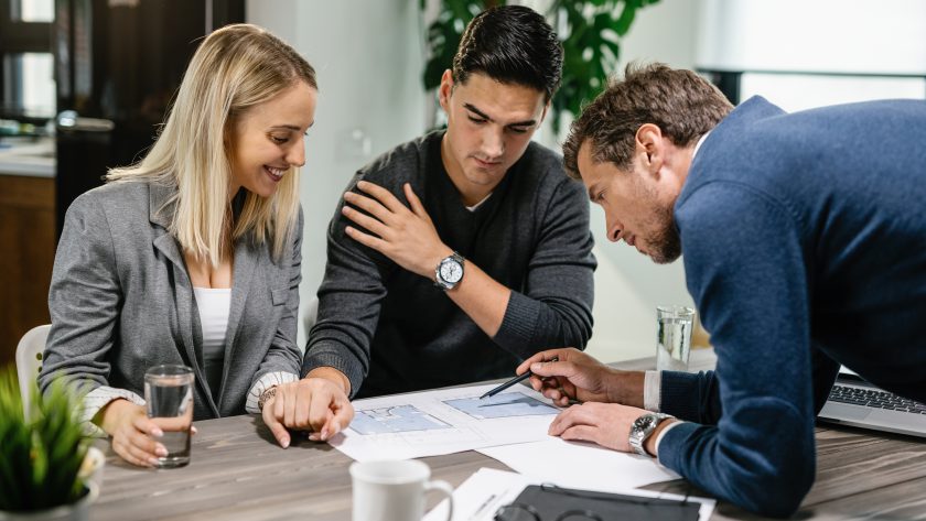 Doradca finansowy wskazuje na dokumenty i omawia plan inwestycyjny z dwojgiem młodych przedsiębiorców przy drewnianym stole.