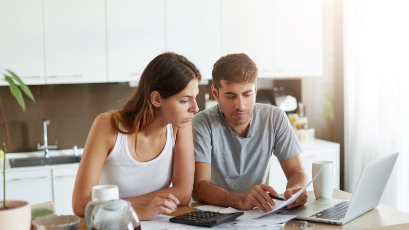 Młoda para siedząca w kuchni przed laptopem, ze skupieniem i troską przeglądająca rachunki oraz dokumenty finansowe.