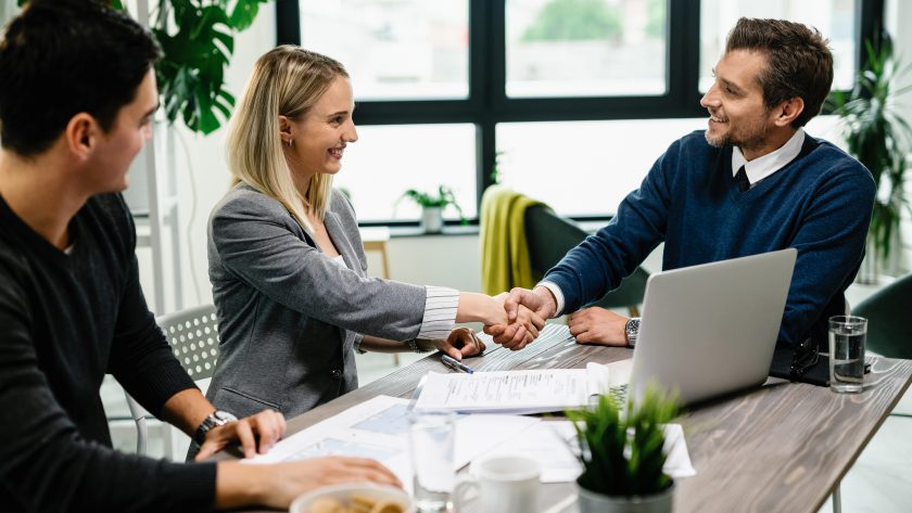 Młoda kobieta w żakiecie i mężczyzna w swetrze ściskają dłoń uśmiechniętego doradcy kredytowego w jasnym biurze z laptopem. Wygląda to na pomyślną rozmowę kredytową.