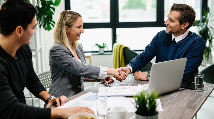 Młoda kobieta w żakiecie i mężczyzna w swetrze ściskają dłoń uśmiechniętego doradcy kredytowego w jasnym biurze z laptopem. Wygląda to na pomyślną rozmowę kredytową.