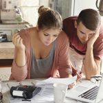 Młoda para siedząca przy kuchennym stole, ze skupieniem i lekkim niepokojem analizująca dokumenty finansowe przed otwartym laptopem.
