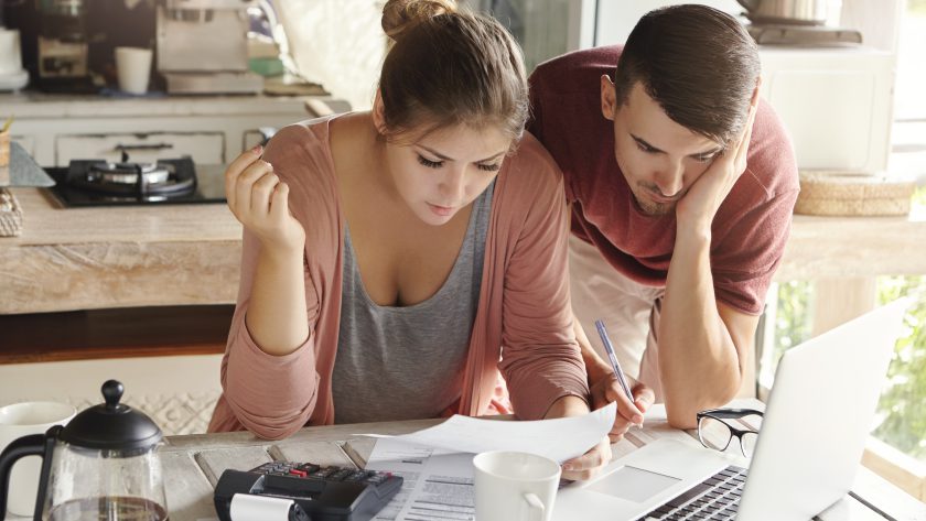 Młoda para siedząca przy kuchennym stole, ze skupieniem i lekkim niepokojem analizująca dokumenty finansowe przed otwartym laptopem.