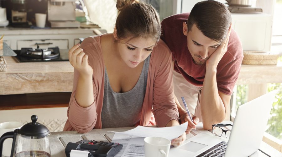 Młoda para siedząca przy kuchennym stole, ze skupieniem i lekkim niepokojem analizująca dokumenty finansowe przed otwartym laptopem.